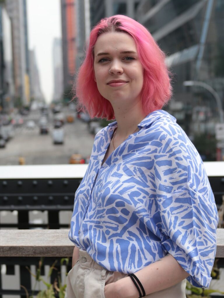 Photo of myself, waist up with a blue and white blouse withpink hair, New York in the Background